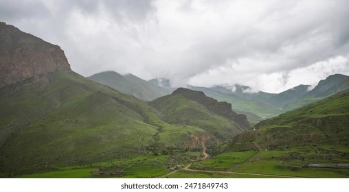 a vast mountainous landscape under an overcast sky. Rolling hills with varying shades of green . A winding road cuts through the valley. Small structures are visible in the distance. - Powered by Shutterstock