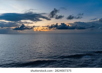 Vast ocean surface under a dramatic sunset sky with dark clouds and warm orange hues. Gentle waves ripple across the water reflecting the fading light, creating a peaceful seascape.