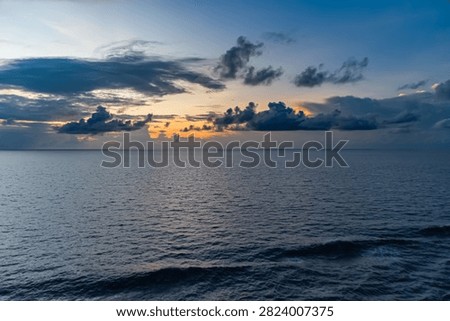 Vast ocean surface under a dramatic sunset sky with dark clouds and warm orange hues. Gentle waves ripple across the water reflecting the fading light, creating a peaceful seascape.