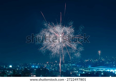 A vast pyrotechnic display with multiple circular explosions and bright trails over a nighttime city with streetlights and buildings in the valley below.