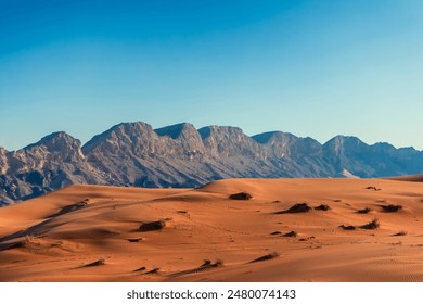 A vast, red sand desert stretches out in front of a range of rocky mountains against a clear blue sky. Photo view of desolation of the desert landscape. Orange sandy desert in Dubai - Powered by Shutterstock