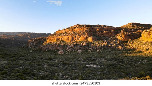 A vast, rugged landscape under a clear blue sky. In the foreground, there is a valley covered in dense shrubs and small trees. In the background, a towering, red rock formation dominates the scene. - Powered by Shutterstock