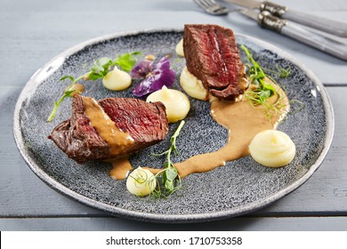 Veal Tenderloin Filet Mignon With Root-crop Cream In Gray Plate. Beef Slices With Sauce, Greenery And Violet Flowers Decoration Close Up. Meat Appetizer, Restaurant Meal, Delicious Dish