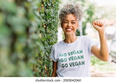 Vegan activist smiling cheerfully while standing alone outdoors. Happy mature woman advocating for veganism while wearing a shirt with the words 