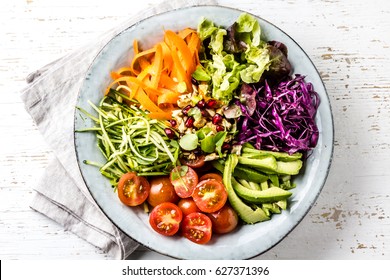 Vegan buddha bowl. Bowl with fresh raw vegetables - cabbage, carrot, zucchini, lettuce, watercress salad, tomatoes cherry and avocado, nuts and pomegranate. White background, top view