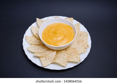 Vegan Cheddar Cheese In A Bowl And Nachos To Dip In A Plate On A Black Background.