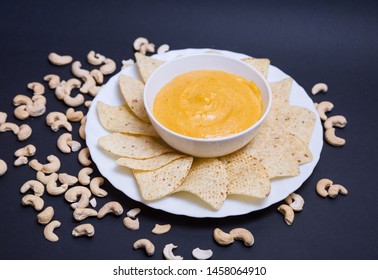 Vegan Cheddar Cheese In A Bowl And Nachos To Dip In A Plate On A Black Background With Cashew Nuts.