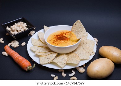 Vegan Cheddar Cheese With Paprika In A Bowl And Nachos To Dip In A Plate On A Black Background With Cashew Nuts, Potatoes And A Carrot.