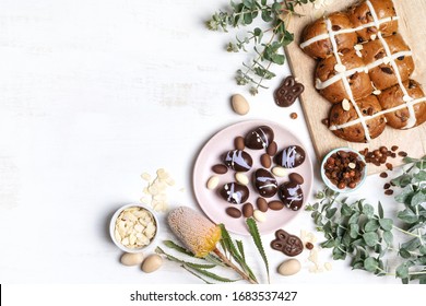 Vegan chocolate Easter bunny, Easter eggs and homemade hot cross buns on a wooden serve board. Decorated by shaved almonds, hazelnuts , Australian Banksia and eucalyptus leaves on a white background.