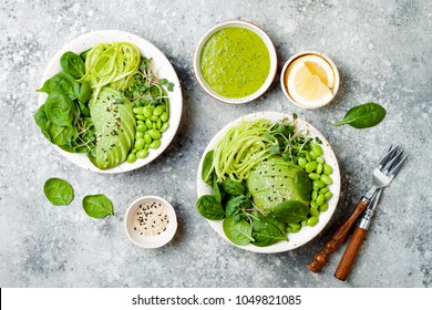 Vegan, detox Buddha bowl with avocado, spinach, micro greens, edamame beans, zucchini noodles and herb green dressing. Top view, grey concrete background