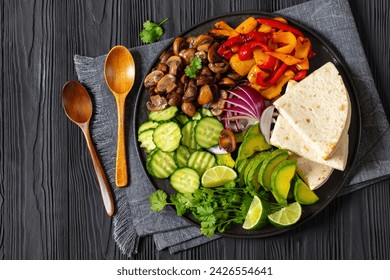 vegan fajitas, mushrooms, bell pepper, red onion, cucumber, avocado, cilantro, tortillas on black plate on black  wooden table with spoons, horizontal view from above, flat lay, free space - Powered by Shutterstock