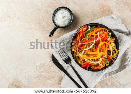 Vegan food, diet. Salad from raw vegetable noodles, pasta from carrot, zucchini, bell pepper. In portioned black bowl with yogurt sauce on a stone table. Top view copy space