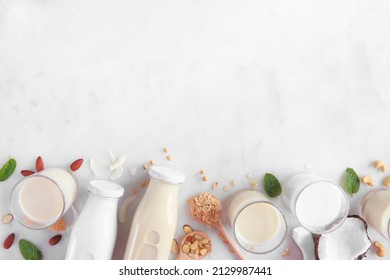Vegan, plant based, non dairy milk bottom border. Variety of types in milk bottles and glasses with scattered ingredients. Top view over a white marble background with copy space.