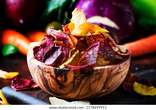Vegan snacks, multicolored vegetable chips in\
wooden bowl and set of fresh farmer vegetables, rustic still life,\
selective focus