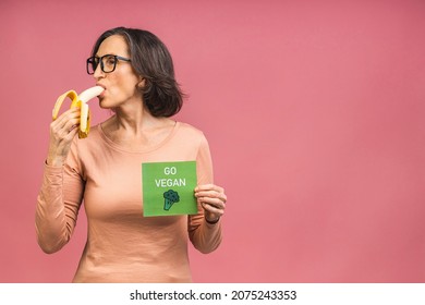 Vegan Or Vegetarian Concept. Aged Mature Senior Woman In Casual Eating Banana. Isolated Over Pink Background.