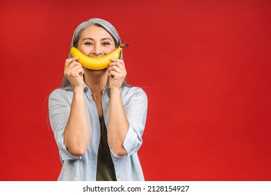 Vegan Or Vegetarian Concept. Portrait Of A Beautiful Elderly Asian Mature Aged Woman Holding Banana Fruit, Smiling, Isolated Over Red Background.