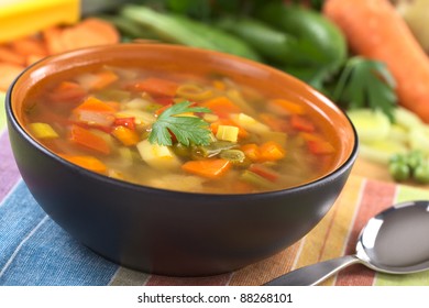 Vegetable soup made of green bean, pea, carrot, potato, red bell pepper, tomato and leek in black bowl garnished with a parsley leaf (Selective Focus, Focus on the front of the parsley leaf)
