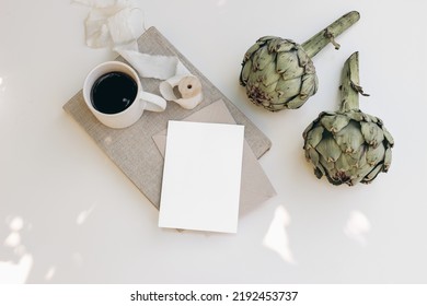 Vegetable Stationery Still Life Scene. Cup Of Coffee, Green Artichoke And Blank Greeting Card Mockup In Sunlight. Beige Table Background With Old Book. Dappled Light, Shadows. Flat Lay, Top View.