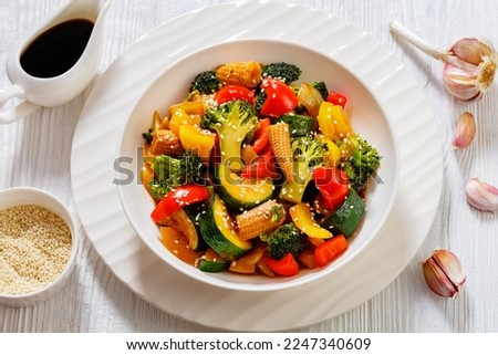 vegetable stir-fry of bell pepper, onion, zucchini, baby corn in cobs, broccoli florets poured with sticky soy sauce in white bowl on white wood table, horizontal view