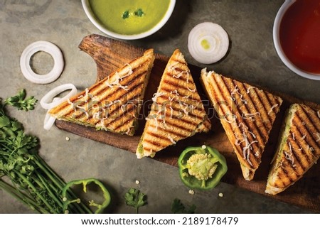 Vegetable Toste sandwich is a type of veg sandwich consisting of a vegetable filling between bread. photo is composited on grey background and wood platter with onion, capsicums  tomato, green sauces.