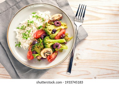 Vegetables like broccoli, tomatoes and olives with rice on a gray plate and a bright wooden table with napkin and fork, healthy vegetarian meal, copy space, high angle view from above, selected 