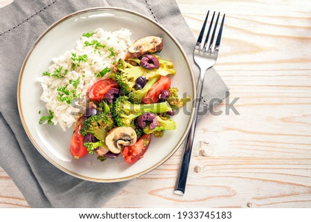 Vegetables like broccoli, tomatoes and olives with rice on a gray plate and a bright wooden table with napkin and fork, healthy vegetarian meal, copy space, high angle view from above, selected 