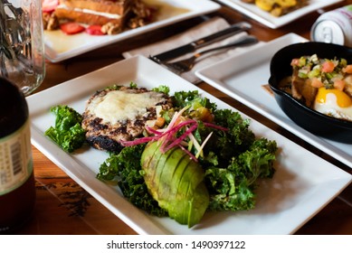 Vegetarian Black Bean Burger With No Bun And Avacado In Foreground On White Plate Around Other Food