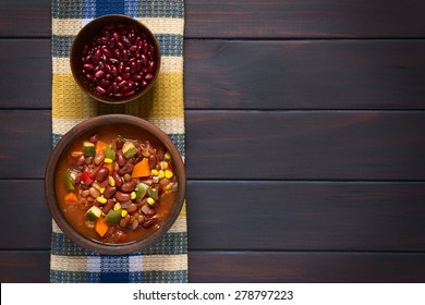 Vegetarian Chili Dish Made Of Kidney Bean, Carrot, Zucchini, Bell Pepper, Sweet Corn, Tomato, Onion, Garlic In Bowl, Raw Kidney Beans Above, Photographed Overhead On Dark Wood With Natural Light