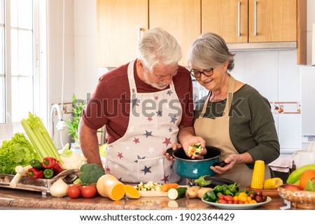 Vegetarian lifestyle. Beautiful white-haired senior couple in the kitchen prepare a vegetable soup. The chef puts chopped vegetables in the pot, on the table a mix of raw seasonal vegetables