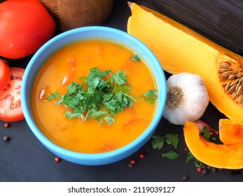 Vegetarian pumpkin soup in a blue bowl. Fresh tomatoes, garlic, pumpkin pieces, wooden utensils on a dark vintage background
