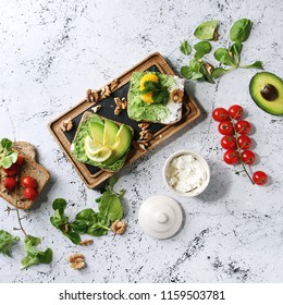 Vegetarian sandwiches with avocado, ricotta, egg yolk, spinach, tomatoes on whole grain toast bread on wooden slate board. Iingredients above, white marble background. Flat lay. Square image
