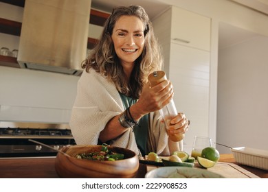 Vegetarian woman smiling at the camera while preparing green juice. Happy woman serving herself a plant-based meal at home. Mature woman taking care of her aging body with a healthy vegan diet. - Powered by Shutterstock