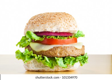 A Veggie Burger (made From Soya Protein) In A Sesame Seed Bap With Layers Of Curly Lettuce, Melted Cheese, Tomato And Onion, On A Light Wood Table With White Background.