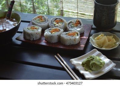 Veggie Sushi Roll With Carrot, Cucumber, Avacado On Sushi Plate, Small Plates Ginger Slices And Green Wasabi Paste, Wood Chopsticks, Red Miso Soup On Black Table Illuminated Through Matchstick Blind