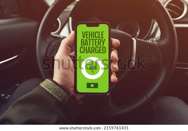 Vehicle battery\
charged at full 100% message on smartphone app screen held by\
electric car driver, selective\
focus