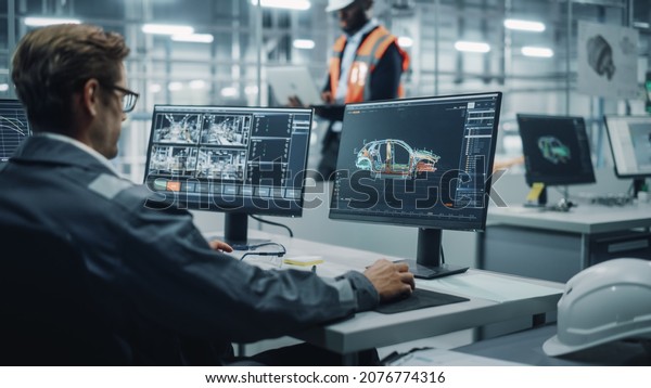 Vehicle Factory Line Operator Working at Desk,\
Overviewing Autonomous Electric Car Production. In the Background\
African American Engineer Using Tablet and Looking at a Car\
Assembly Plant.