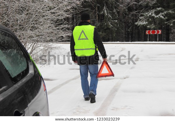 Vehicle safety at a stop, a traffic accident - a man\
driver in a black jacket and a bright yellow fluorescent signal\
vest  goes to put up an emergency stop sign - red triangle on a\
forest snow road