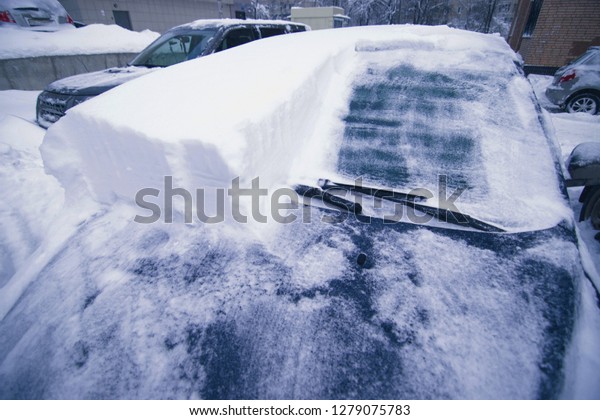 Vehicles covered with snow in the winter blizzard\
in the parking. Cars in snowdrifts after a snowfall. After the\
winter storm
