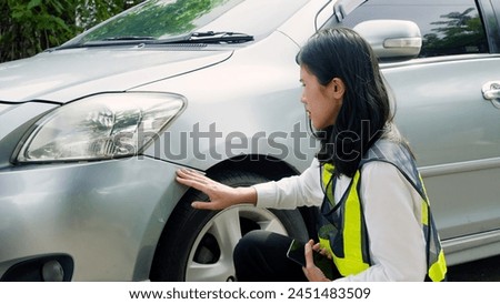 Veiled woman in safety vest, crouching by car, inspecting, holding phone, outdoor setting. Concept of taking care of car insurance after an accident.