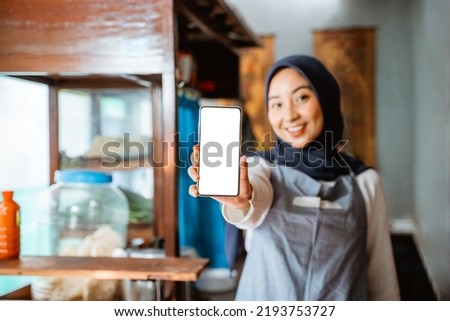 veiled woman wearing apron showing cellphone screen while selling in chicken noodle cart