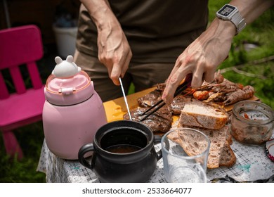 Veiny hands skillfully cut grilled meat using chopsticks and a knife, with fresh bread, a pink thermos, and a black clay dish. The scene captures the essence of outdoor dining. - Powered by Shutterstock