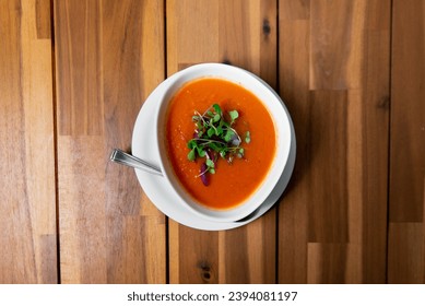 Velvety tomato soup topped with fresh microgreens, served in a white bowl against a warm wooden backdrop, captured from above for a comforting visual feast - Powered by Shutterstock