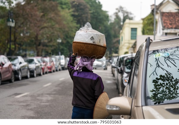 Vendor with basket of bread on head on Hanoi\
street, Vietnam. Two rows of cars parking on street. Concept of\
contrast between living\
level