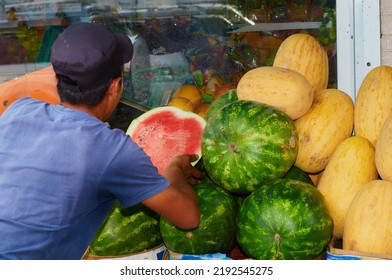 A Vendor Lays Out Half A Red Watermelon On An Open-air Farmers' Market Stall. Stacked Large Watermelons And Melons. The Juicy Pulp Of The Watermelon Is Protected By A Transparent Plastic Cling Film.