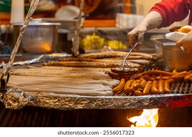 A vendor tends to a large grill filled with various types of sausages and hot dogs at a bustling local market as evening approaches, filling the air with delicious aromas. - Powered by Shutterstock