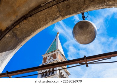 Venetian bell tower of St. Mark's Basilica in Venice against the blue sky from an unusual perspective, the top of the tower with an angel shining in the sun framed by a stone arch, Italian landmarks