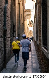 Venice, Italy - April 20, 2019: Man And Woman With Matching Captain's Hats Walking Along A Narrow Dark Alley With Shabby Brick Buildings Towards A Group Of People Standing In The Light