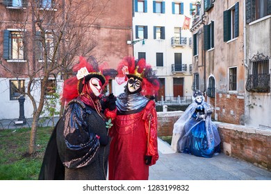 Venice, Italy - February 10, 2018: Couple In Queen Of Hearts And King Of Clubs Black And Red Costumes And White Masks And The Woman Sitting In The Background On The Street During The Carnival