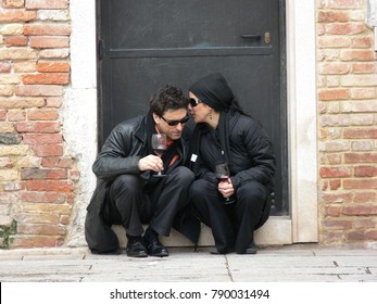 Venice, Italy - February 25 2006: Attractive Black Dressed Couple With Sunglasses Sitting On A Threshold Of An Old House, Drinking Wine And Whispering
