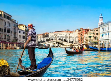 Venice, Italy. Gondolier dressed in traditional blue striped top and straw hat with red ribbon, with rowing oar in his gondola on Grand Canal look at Rialto Bridge against other gondolas in sunny day